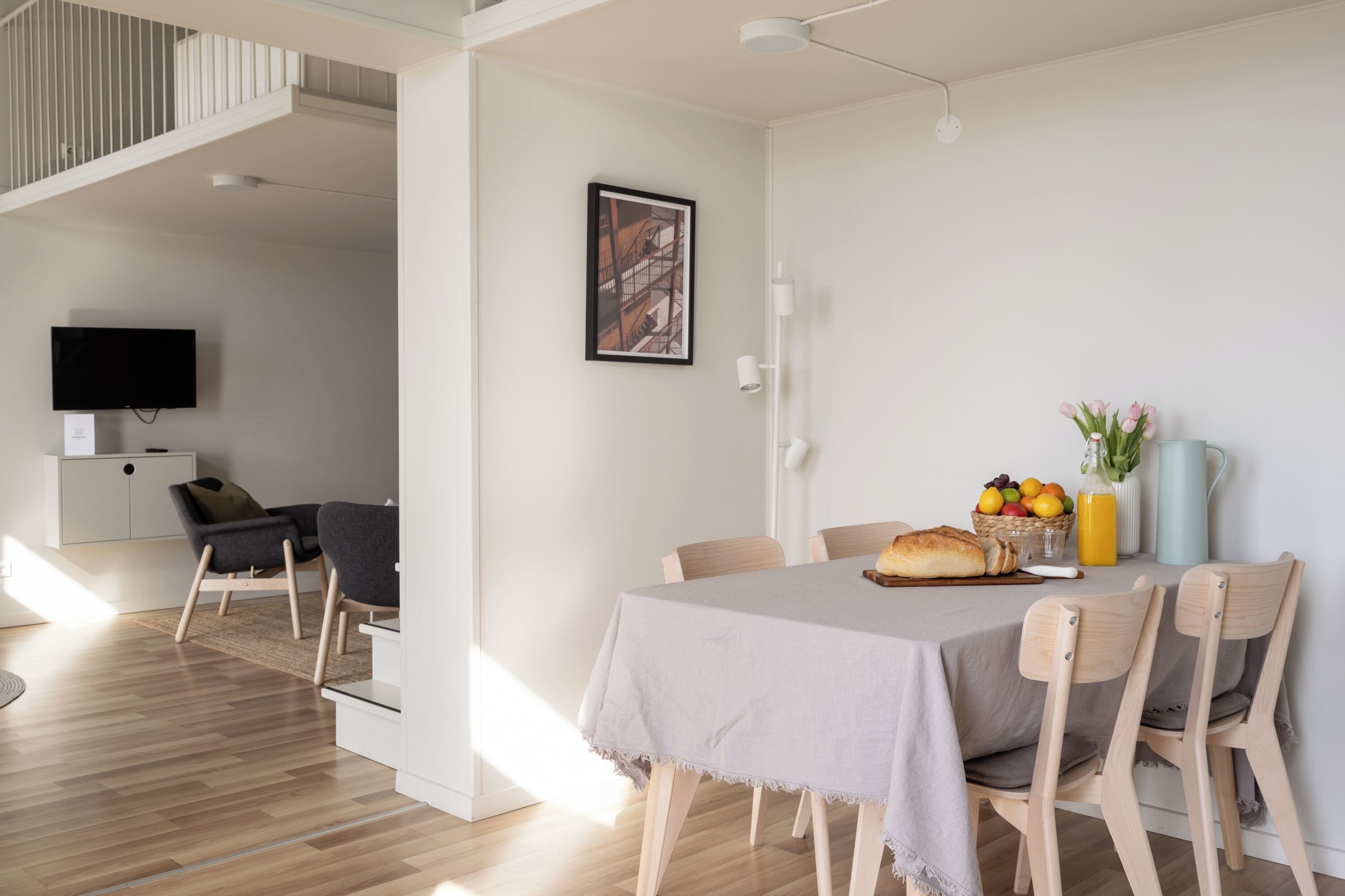 A modern dining area with a table set for four, a television mounted on the wall, and a staircase leading to an upper level.