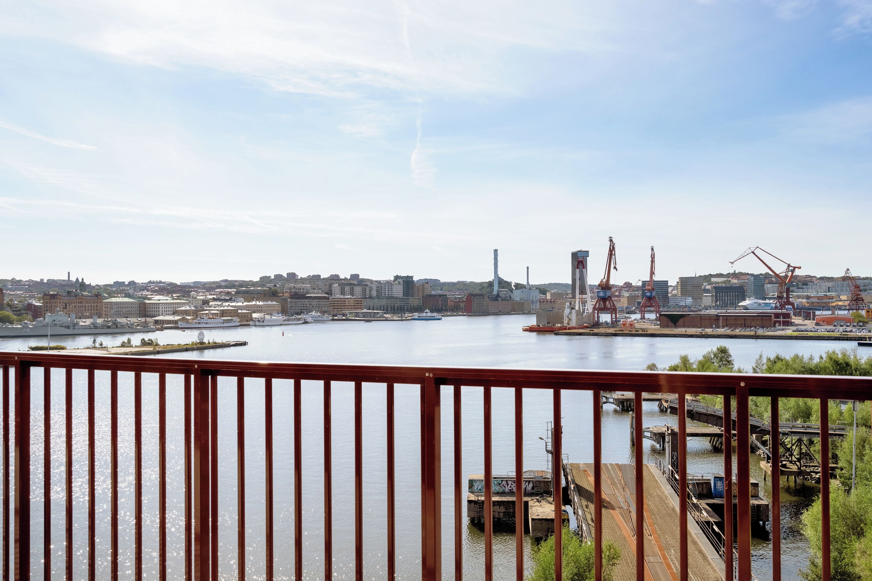 A waterfront view with industrial buildings and cranes, a calm river, and a metal railing in the foreground.