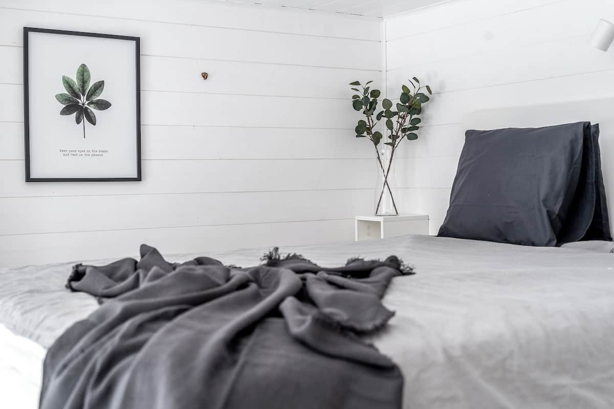 Un dormitorio minimalista con una cama, un grabado botánico enmarco, una planta en maceta y una mesita de noche.