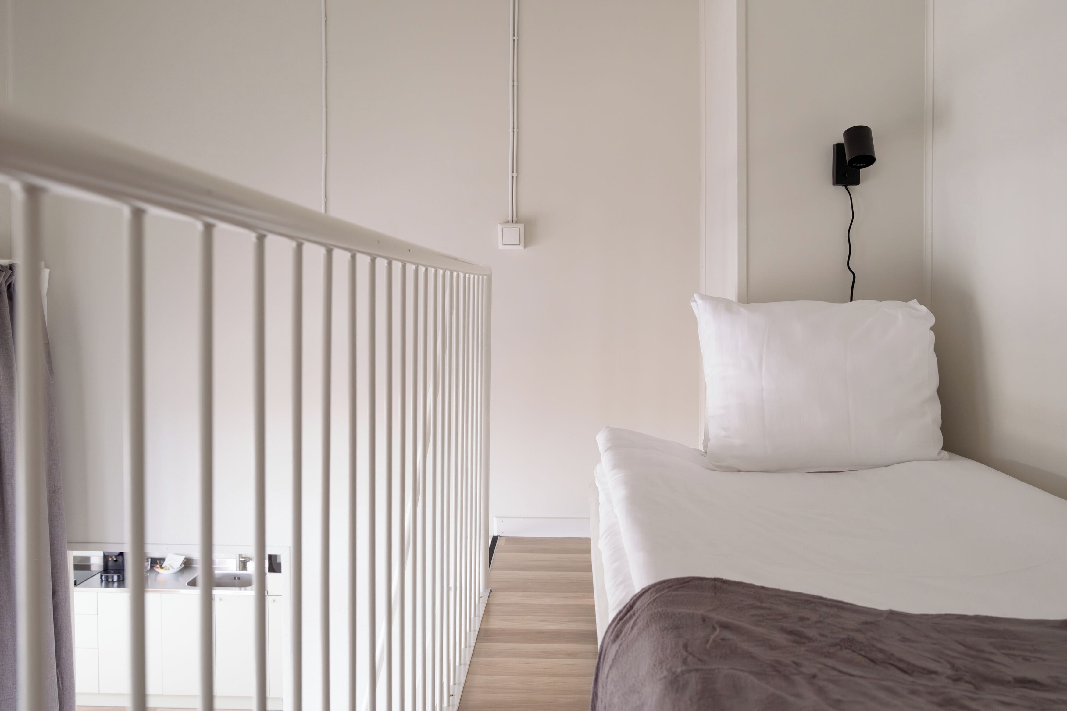 Un dormitorio con una cama, un radiador blanco, una lámpara de pared negra y suelo de madera.