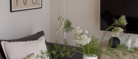 Un salón moderno con una mesa redonda blanca, un jarrón blanco con flores, un sofá gris y un televisor negro en la pared.