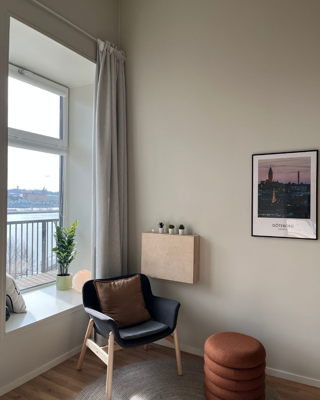 A modern living room with a view of a cityscape, a chair, a small table with plants, and a framed picture on the wall.