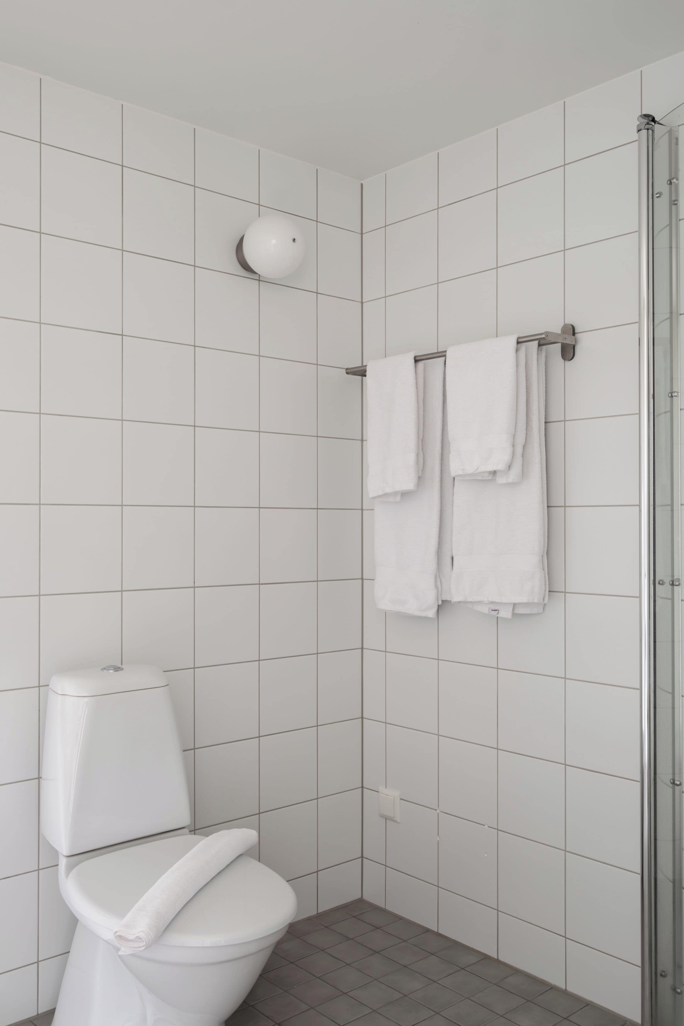 Ett badrum med vita kakelplattor, en toalett, en dusch med en glasdörr och två handdukar som hänger på en stång.