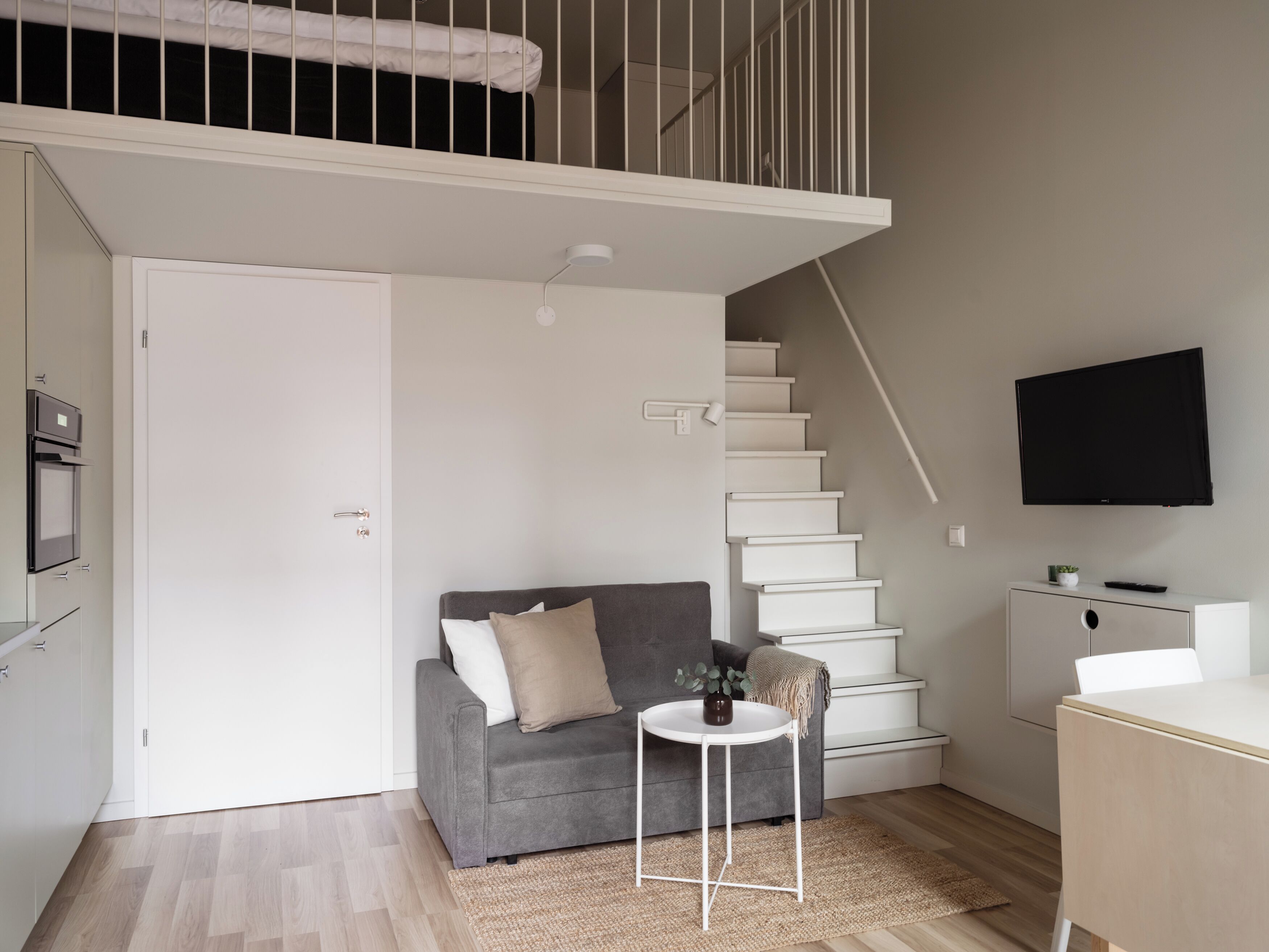 A modern living room with a grey sofa, a small round table, a staircase, and a TV mounted on the wall.
