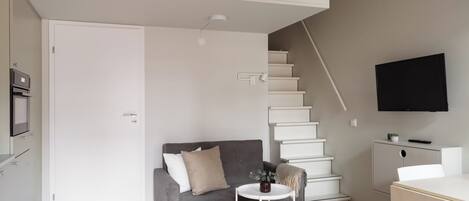 A modern living room with a grey sofa, a small round table, a staircase, and a TV mounted on the wall.