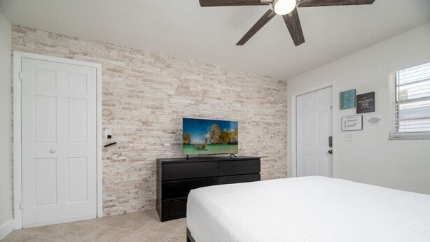 A bedroom with a white brick wall, a ceiling fan, a bed, a dresser, and a TV on the wall. - The Heart of Wilton #5 (Wilton Manors)