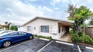 A single-story building with a covered walkway, a parking lot, and a blue car parked in front. - The Heart of Wilton #5 (Wilton Manors)