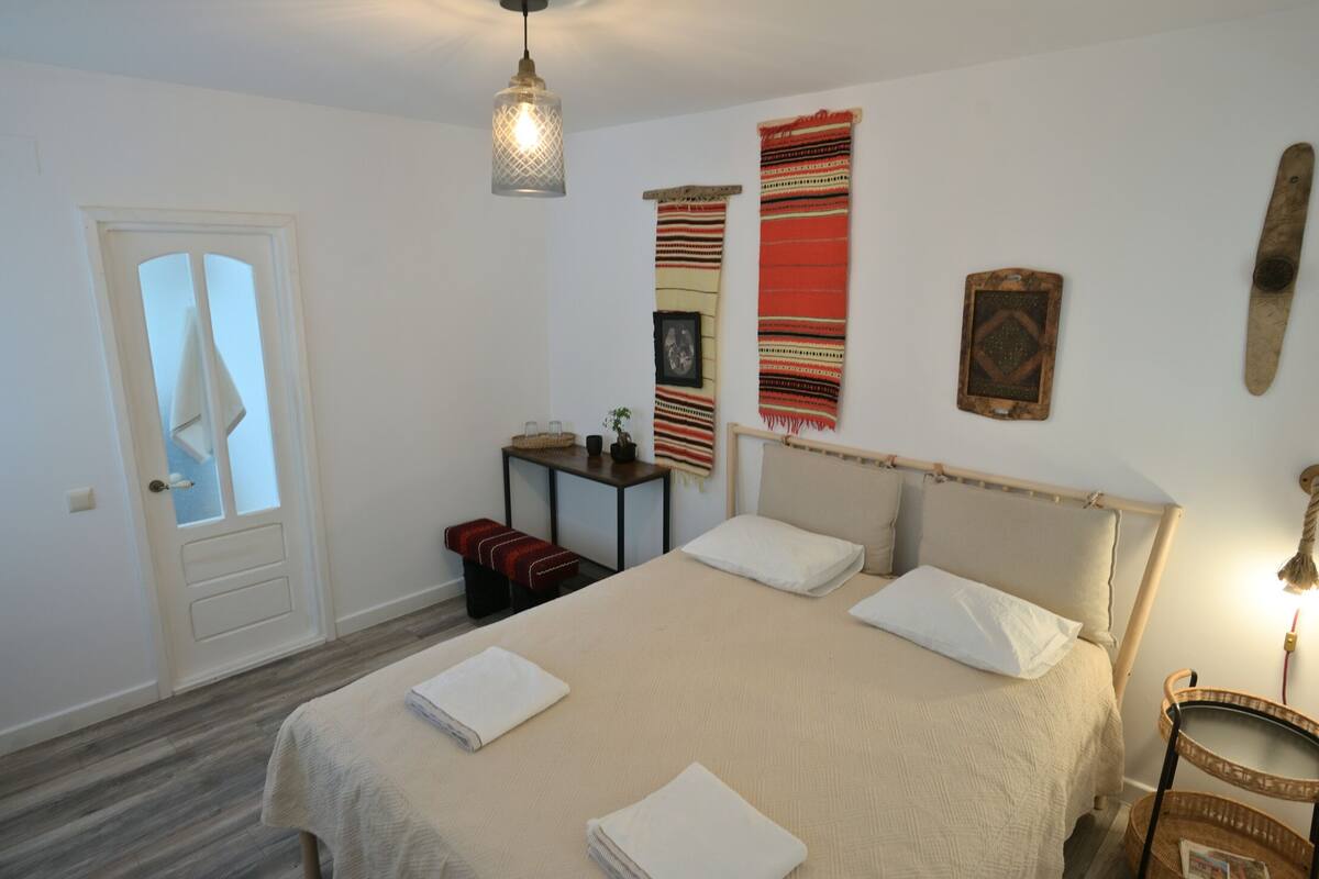 A bedroom with a bed, a bedside table, a hanging light fixture, a door, and a wall with a tapestry.