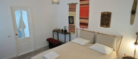 A bedroom with a bed, a bedside table, a hanging light fixture, a door, and a wall with a tapestry.