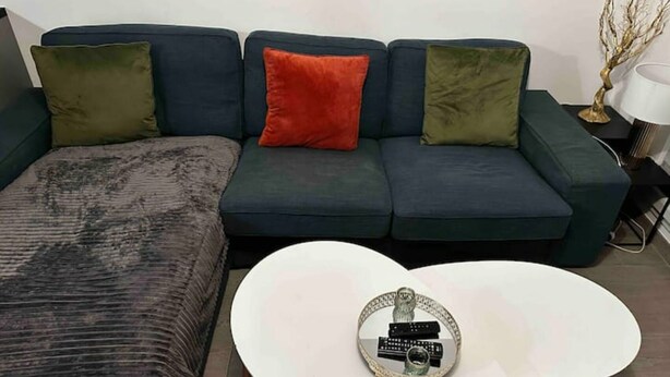 Un salon avec un canapé bleu foncé, des coussins verts et rouges, un pouf à motifs et une table basse blanche sur laquelle reposent des télécommandes.