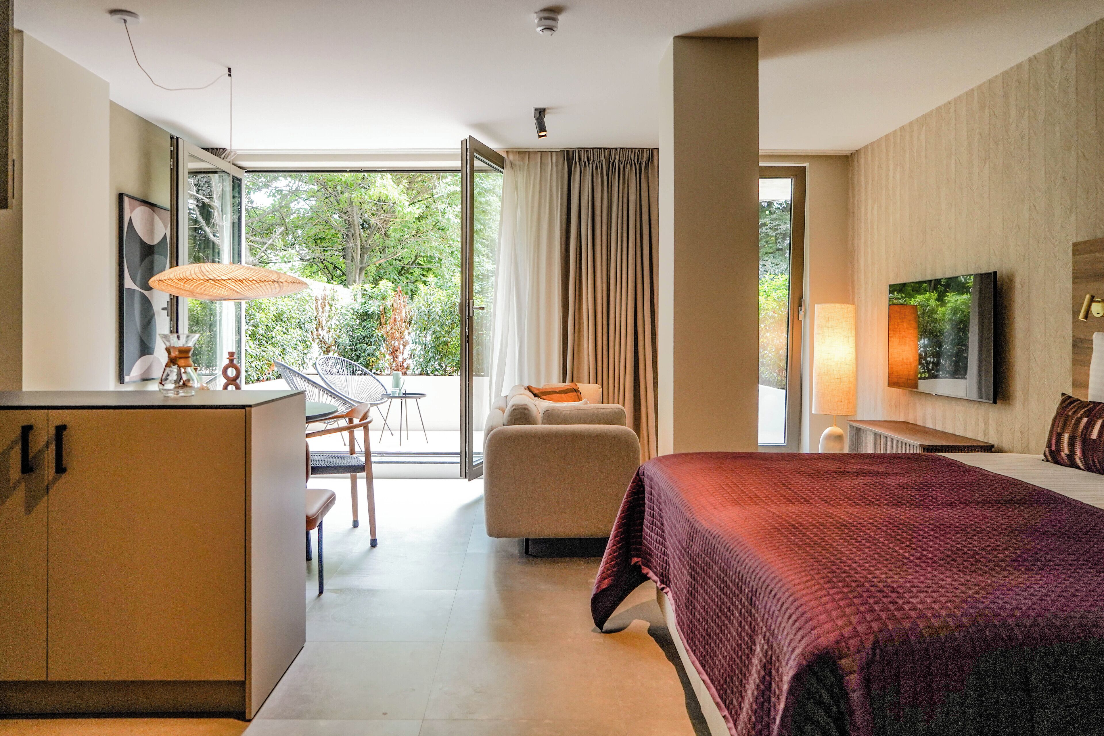 A modern hotel room with a large bed, a desk, a chair, and a balcony with a view of greenery.