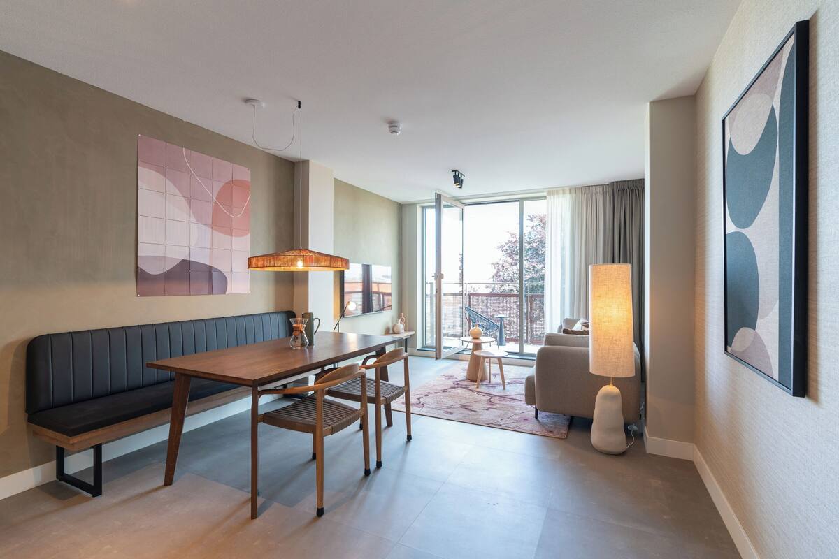 A modern hotel room with a wooden dining table, two chairs, a bench, a large window with curtains, a floor lamp, and abstract art on the walls.