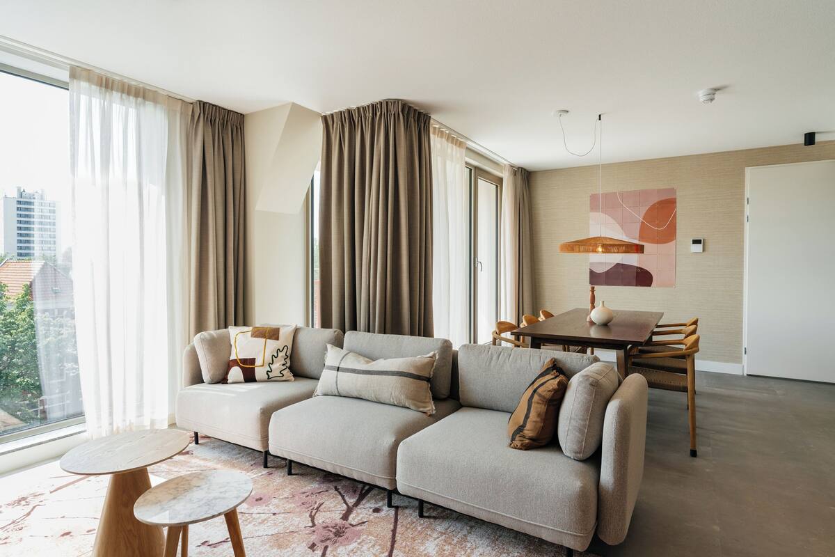 A modern living room with a sectional sofa, a dining table, and a large window with curtains.