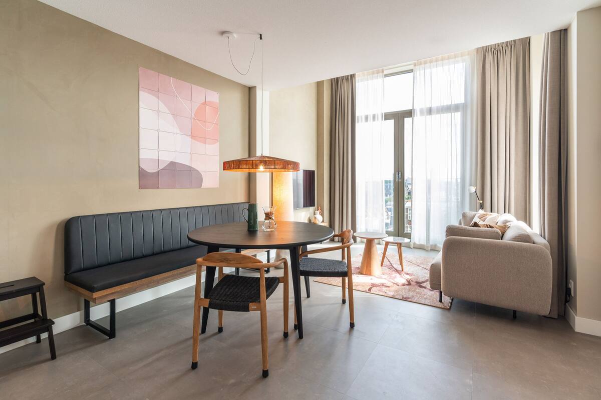A modern living room with a sofa, a round dining table with chairs, a bench, and a large window with curtains.