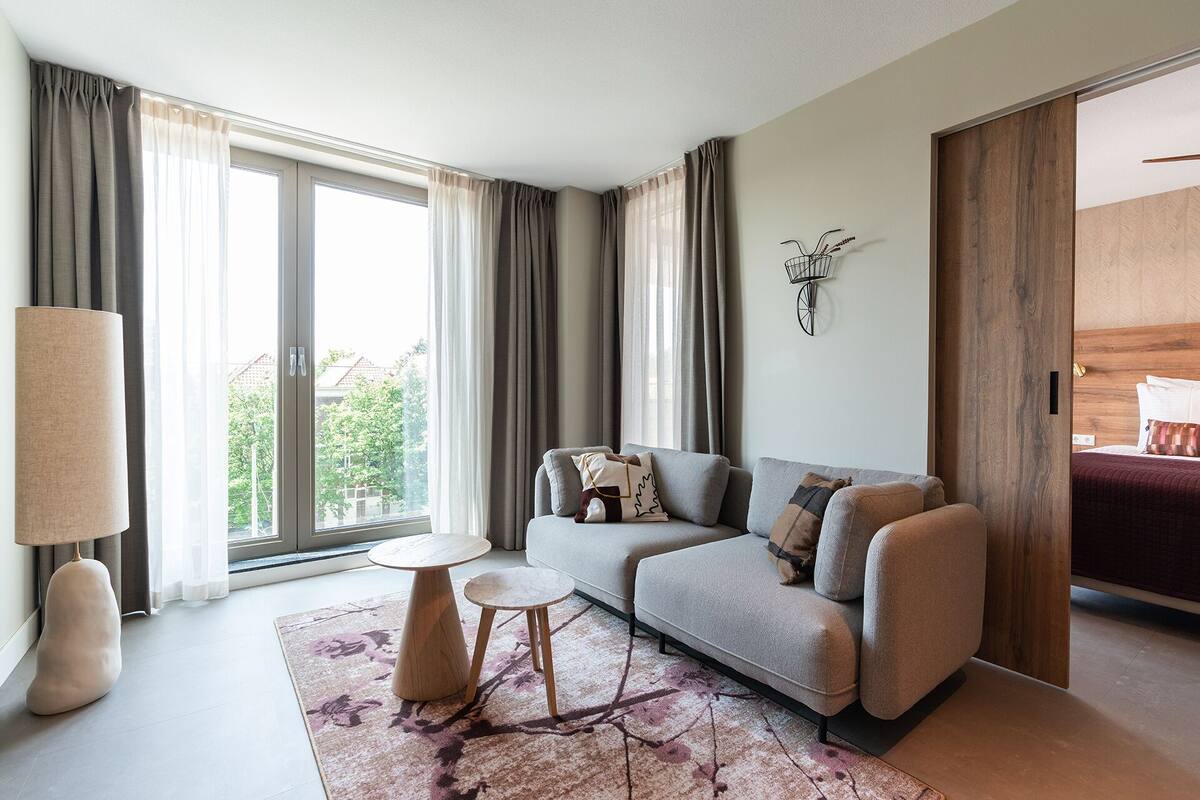 A modern living room with a grey sofa, two small tables, a large floor lamp, and a view of greenery through large windows.