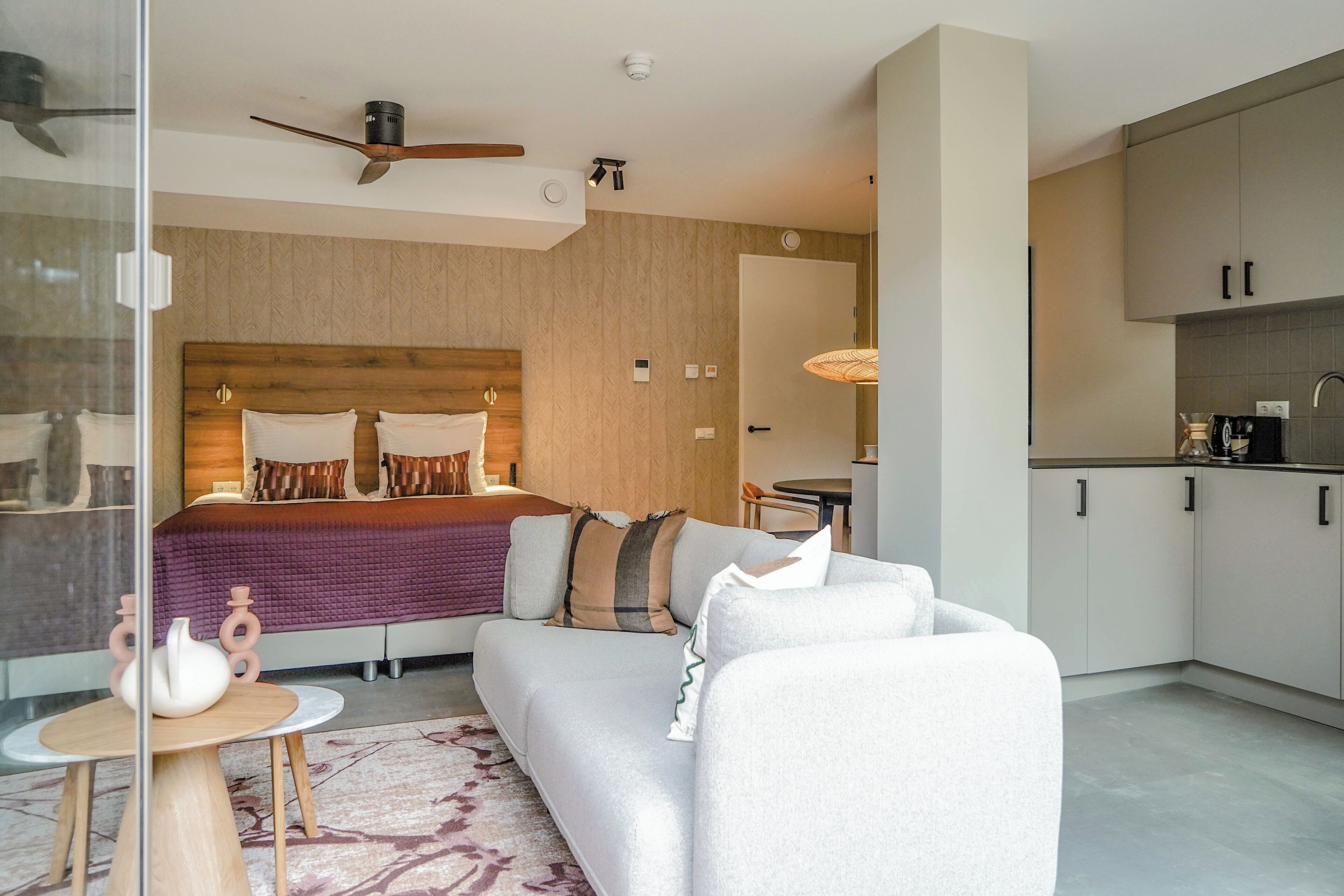 Una habitación de hotel moderna con una cama grande, un sofá, una mesita y una zona de cocina.