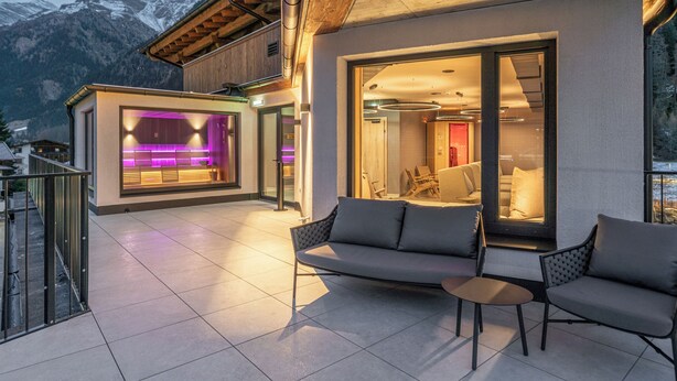 Un espacio exterior moderno con sofá y sillones, una cocina con cerramiento de vidrio y luz púrpura, y montañas al fondo.