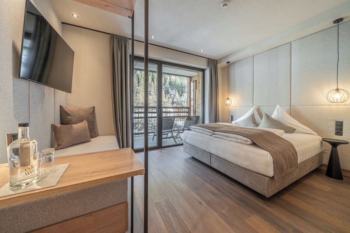A modern bedroom with a large bed, a wooden desk, a TV, and a balcony with a view of trees.