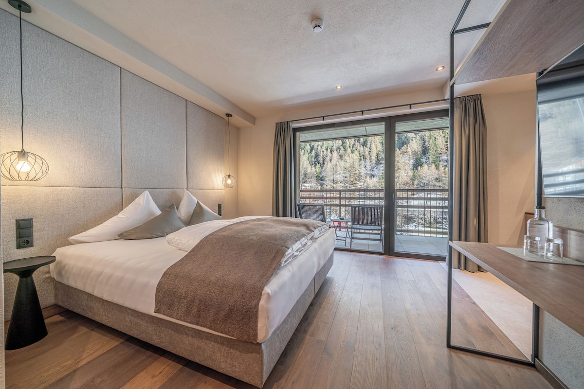 A modern hotel room with a large bed, a small table, and a balcony with a view of trees.