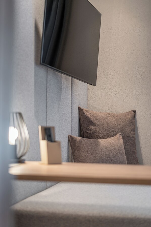 A modern bedroom with a flat-screen TV mounted on the wall, a bedside table with a lamp, and two pillows.