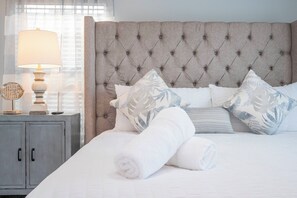 A neatly made bed with white bedding and rolled towels, a tufted headboard, and a bedside table with a lamp and decorative items.
