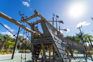 Une structure d’escalade en bois, dotée de cordes et d’un toboggan, entourée de verdure et de palmiers.