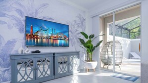 A modern living room with a large TV mounted on the wall, a grey console table, a potted plant, and a glass door leading to an outdoor area.