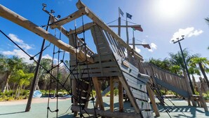 A wooden play structure with ropes and a slide, surrounded by greenery.