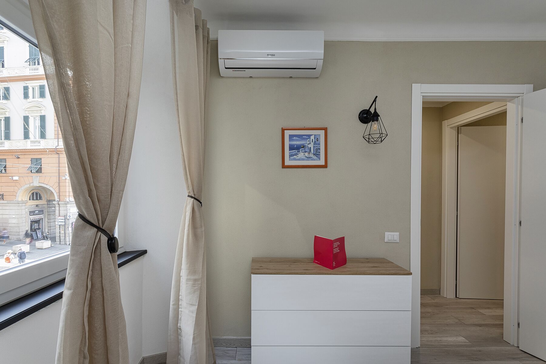 Ein Zimmer mit Fenster, Klimaanlage, einem gerahmten Bild, einer roten Tasche auf einer Holzkommode und einer Tür zu einem anderen Raum.