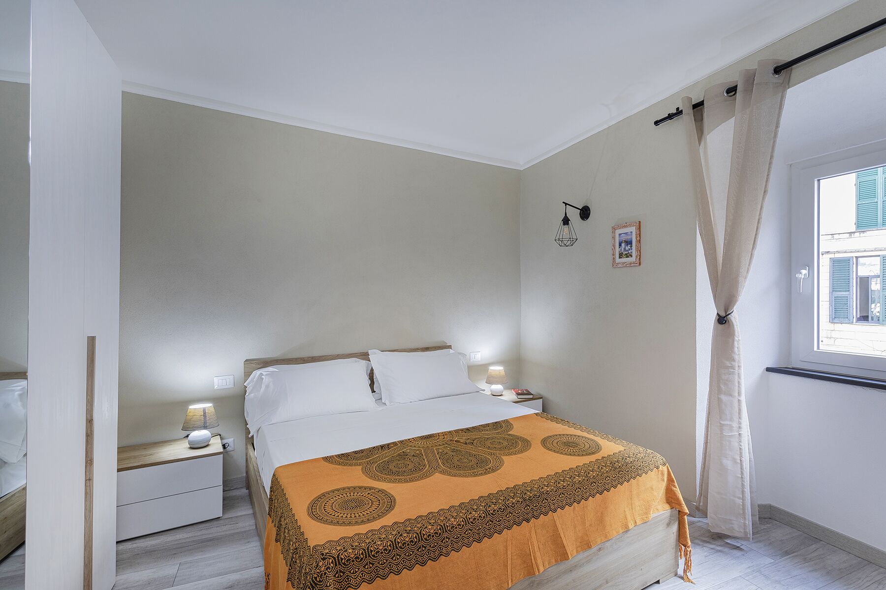 Un dormitorio con cama, mesitas de noche, lámpara, ventana con cortinas y una lámpara de pared.