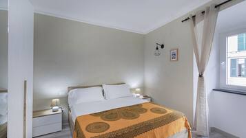 Un dormitorio con cama, mesitas de noche, lámpara, ventana con cortinas y una lámpara de pared.