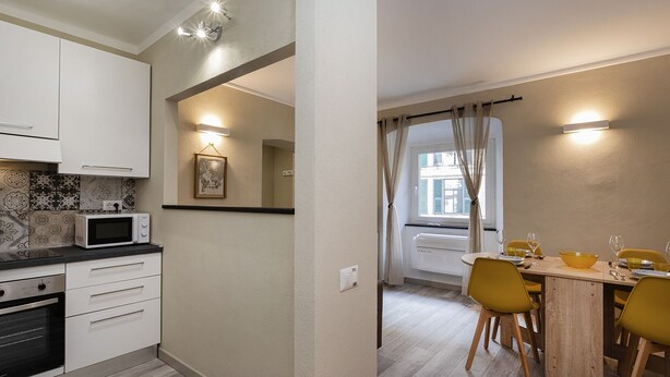 A compact kitchen with white cabinets, a microwave, and a stove. There is a dining area with a table, yellow chairs, and a window with curtains. - Raibetta Apartments by Wonderful Italy (Genoa)