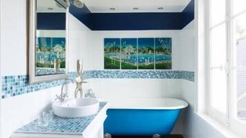 A bathroom with a white vanity, a blue freestanding bathtub, and a window with a view of a house.