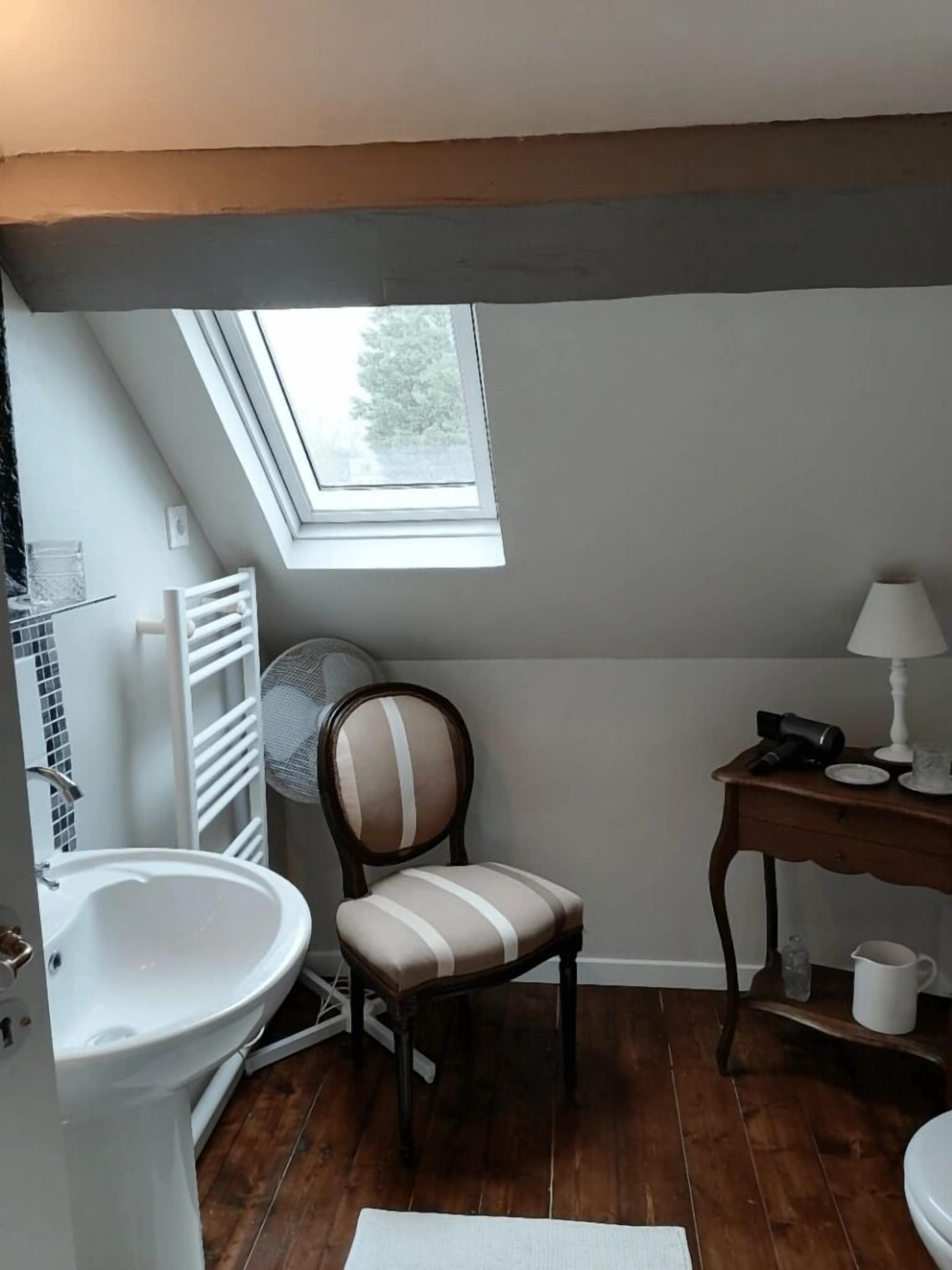 Une petite salle de bain avec un puits de lumière, un lavabo blanc, une chaise et une table en bois sur laquelle se trouve une lampe.