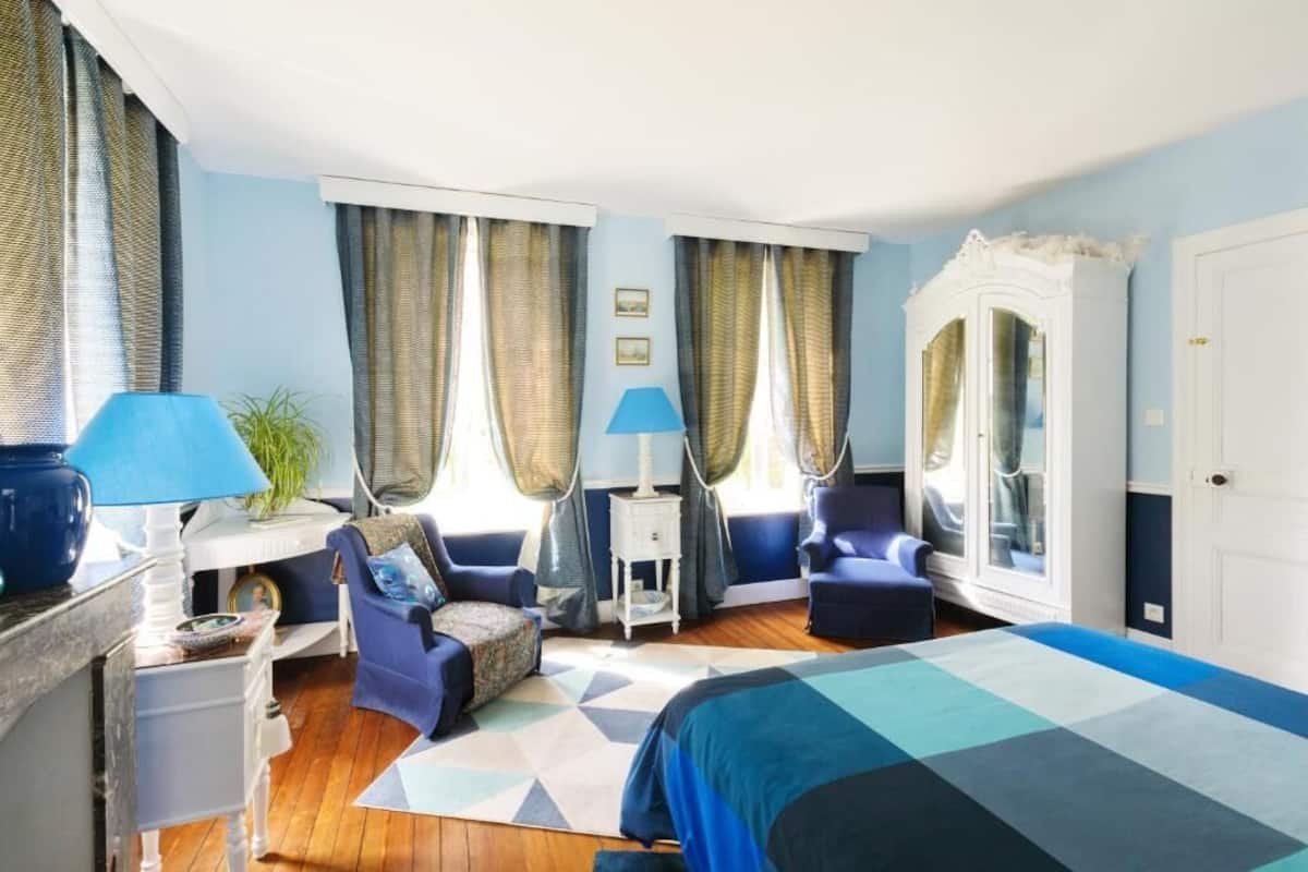 Ein Schlafzimmer mit einem Bett, zwei Stühlen, einem Schreibtisch, einer Lampe und Vorhängen.