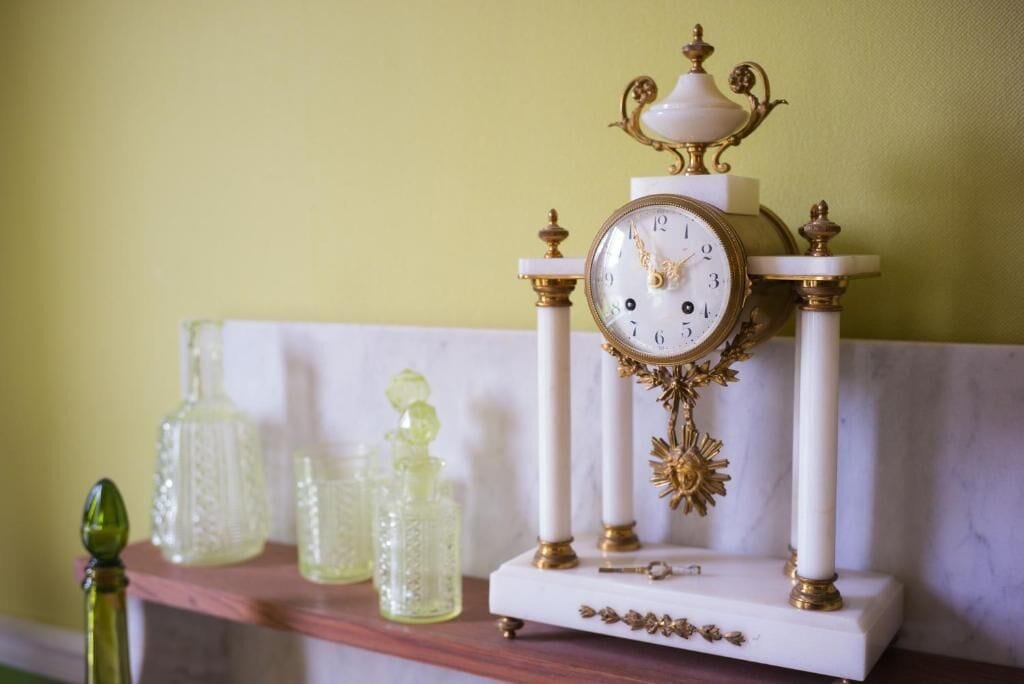 A white and gold ornate clock on a marble pedestal, accompanied by three glass bottles on a shelf.