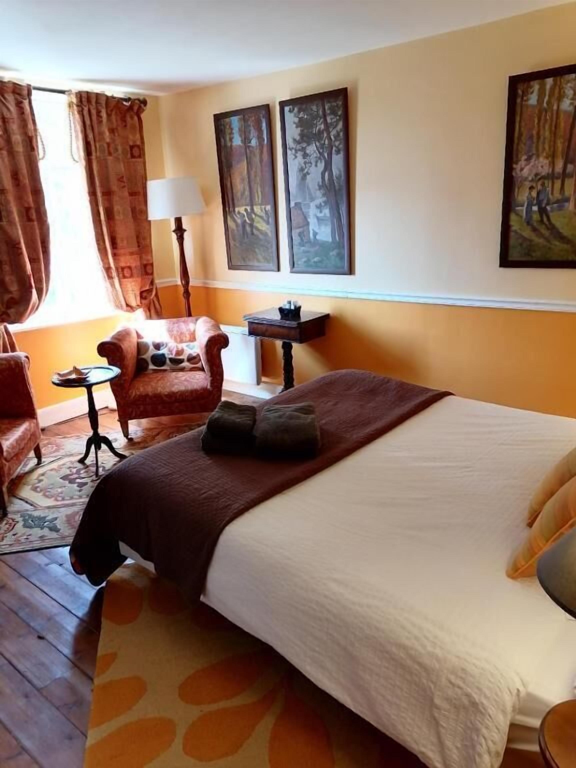 A hotel room with a bed, a chair, a small table, and two paintings on the wall.