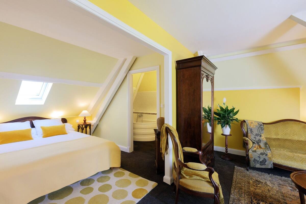 Ein Hotelzimmer mit Bett, Sessel, Sofa, einem kleinen Tisch, einer Lampe, einem Blumentopf und einer Treppe zu einer oberen Ebene.