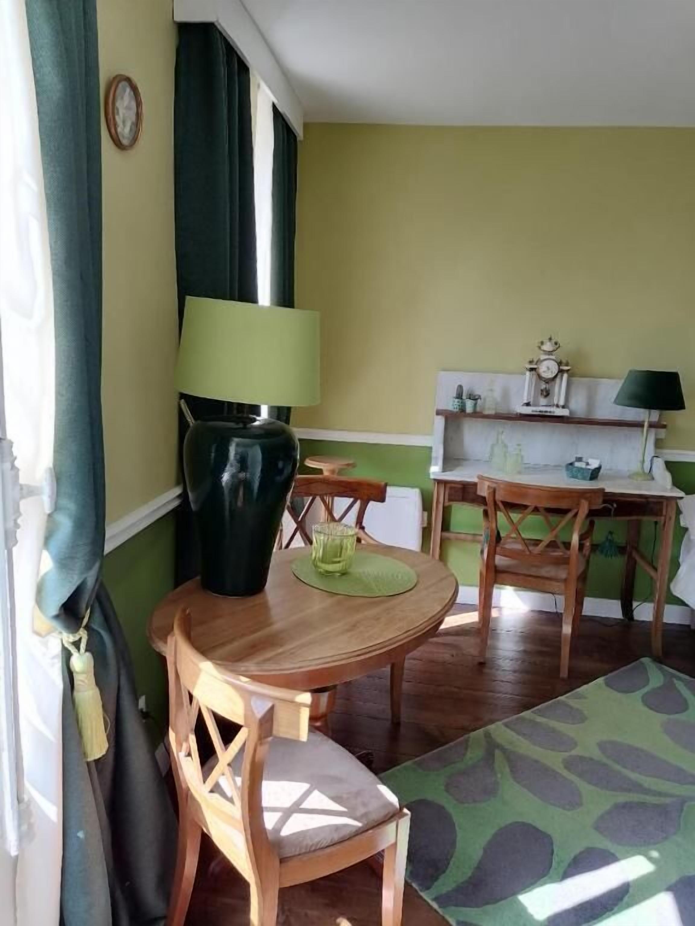 A room with a wooden dining table, chairs, a green patterned rug, and a window with curtains.