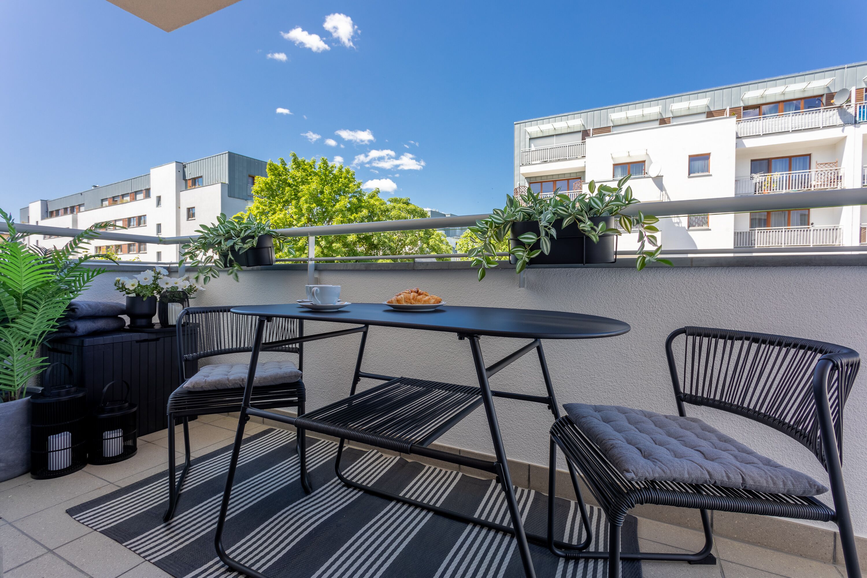 A balcony with a round table and two chairs, potted plants, and a view of a building with balconies.