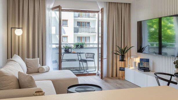 Moderni hotellihuone, jossa on sohva, ruokapöytä, televisio ja parveke, jolta on näkymä rakennukseen.