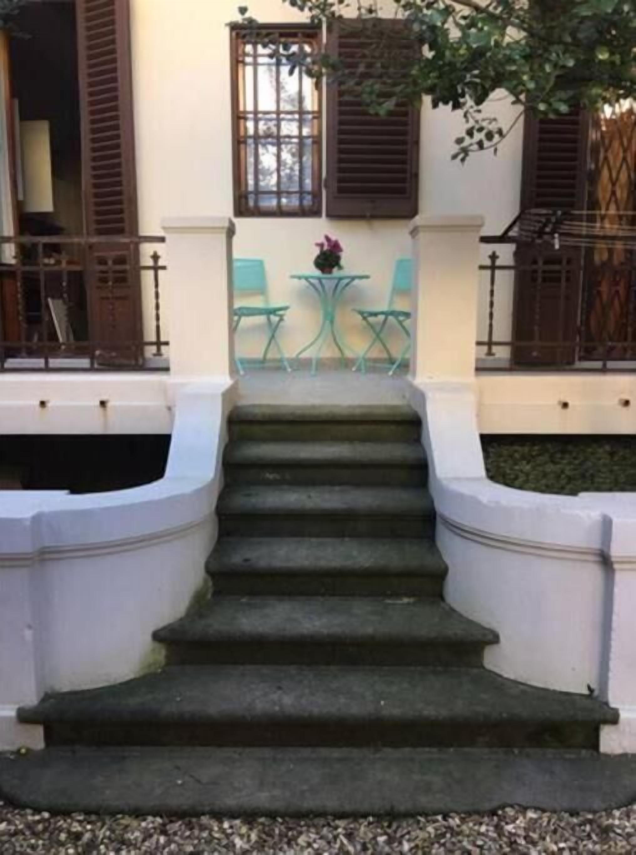Een reeks stenen treden leidt naar een klein terras met twee groene stoelen en een tafel, begrensd door houten leuningen en een raam met luiken.