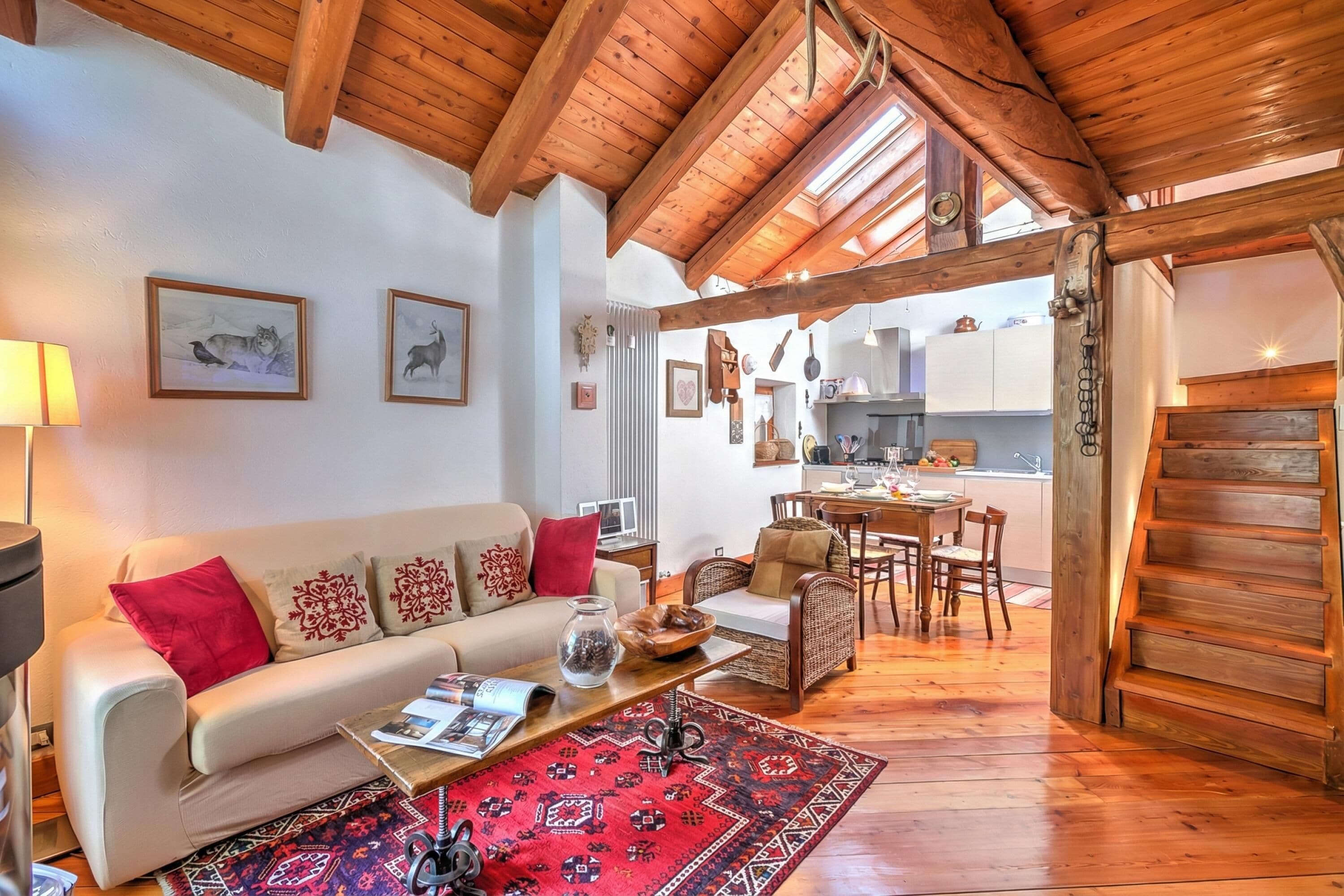 A cozy living room with a wooden ceiling, a sofa with patterned cushions, a coffee table with a magazine, and a red rug.