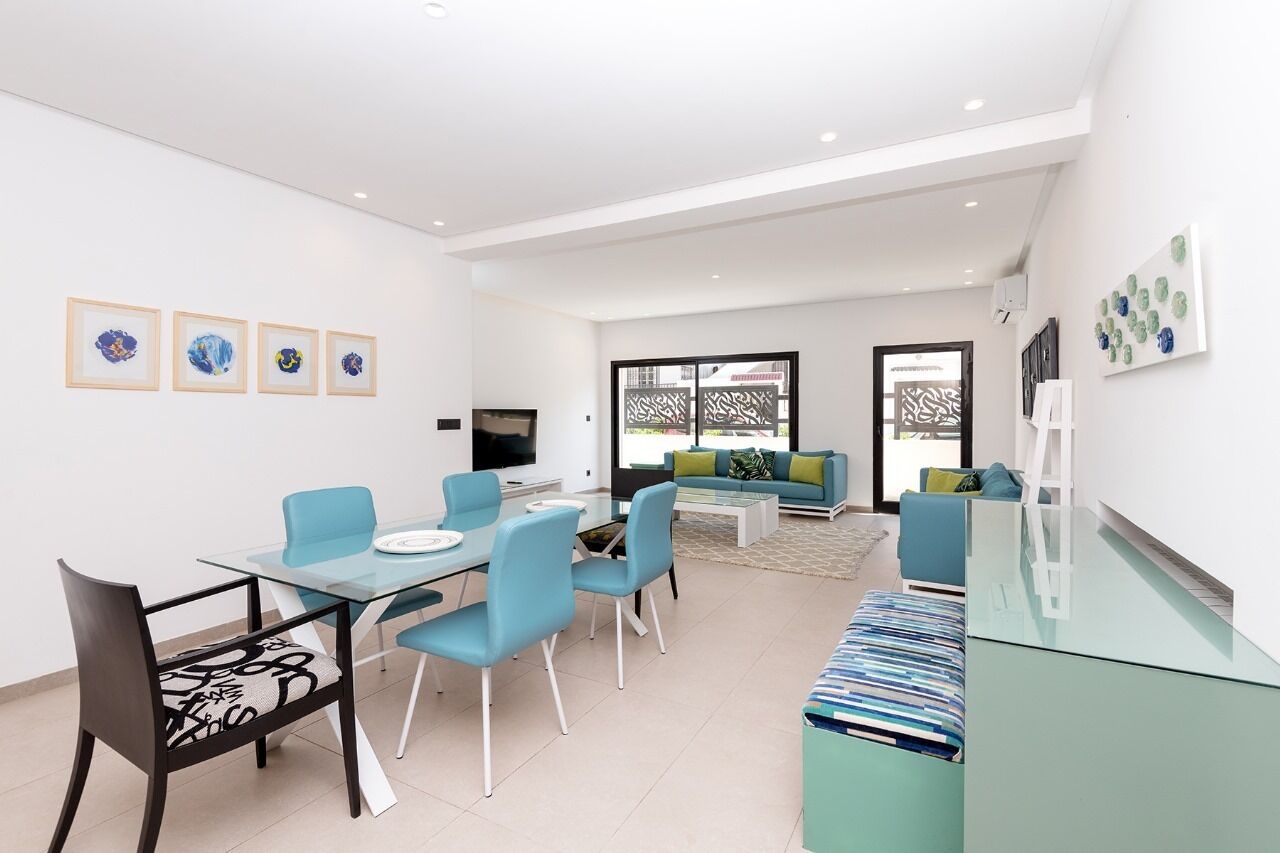 A modern living room with a glass dining table, blue chairs, a sofa, and a TV.