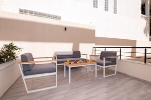 Un espacio exterior moderno con una zona de estar que incluye un sofá y sillas, una mesa de madera con alimentos para el desayuno, y vistas de un edificio blanco con ventanas.