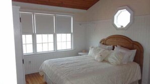 Een slaapkamer met een houten plafond, een groot raam, een bed met wit beddengoed en een klein nachtkastje.