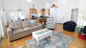A spacious living room with a beige sofa, a white coffee table, and a blue patterned rug. There is a kitchen area with wooden cabinets and a refrigerator.