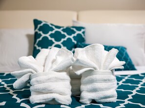A bed with white and blue patterned bedding, two white towels shaped like bunny slippers, and a teal pillow with a white geometric pattern.