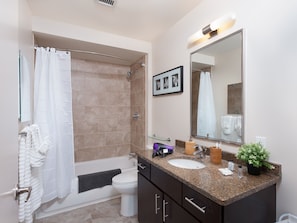 A modern bathroom with a bathtub, toilet, and sink. There is a shower curtain, a framed picture on the wall, and a potted plant on the sink counter.