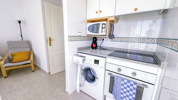 A compact kitchen with a microwave, stove, and washing machine, a chair with a cushion, and a door.
